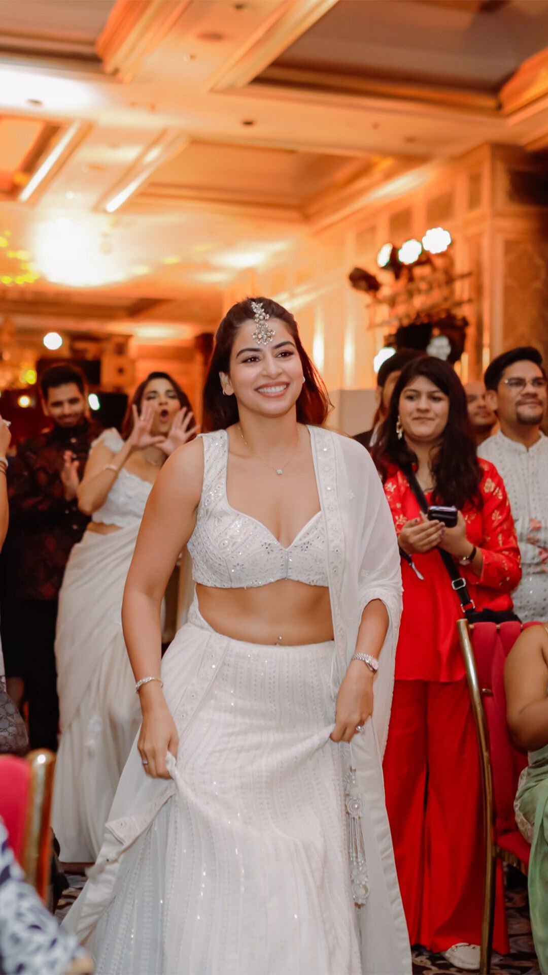 Smiling woman in a white embellished lehenga with matching dupatta and jewelry walking gracefully at an indoor wedding celebration.